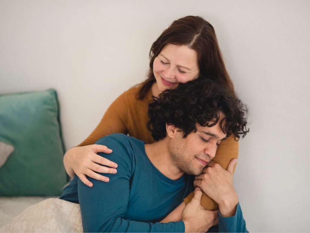 Zwei Menschen sitzen beieinander, eine Person hält die andere im Arm, beide lächeln in einem achtsamen, berührenden Moment.